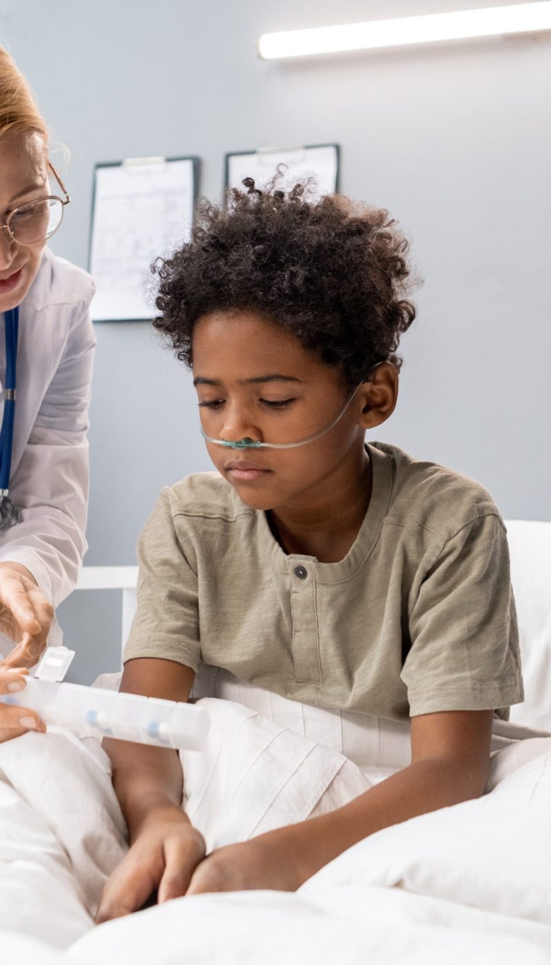 Mature female doctor pointing at medical equipment and explaining the way of treatment to little patient at hospital ward