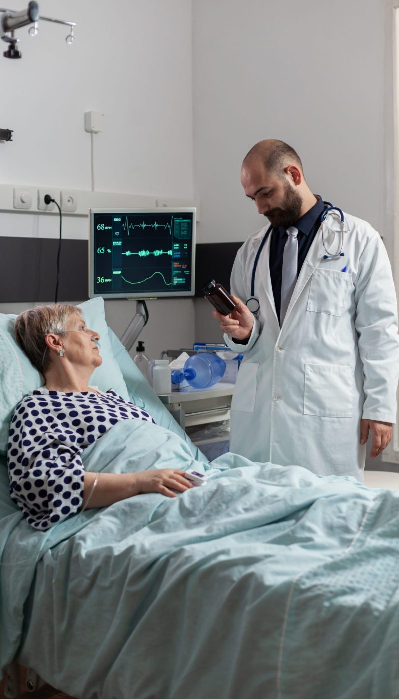 Senior woman patient breathing with help from oxygen tube laying in hospital bed, listening doctor explaying diagnosis treatmant holding pills bottle for recovery.